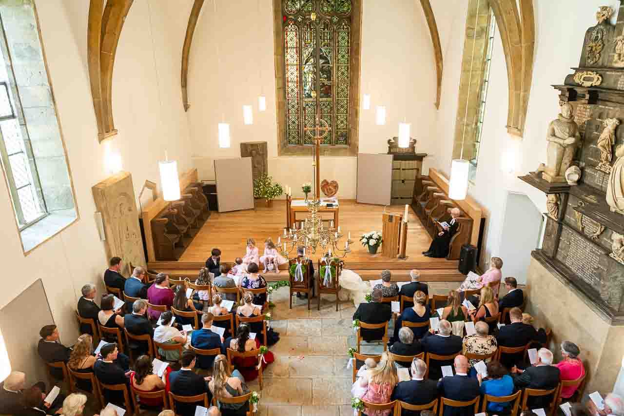 Romantische Trauung in der Kirche: Das Brautpaar gibt sich das Ja-Wort, umgeben von festlich gekleideten Gaesten in historischer Atmosphaere.