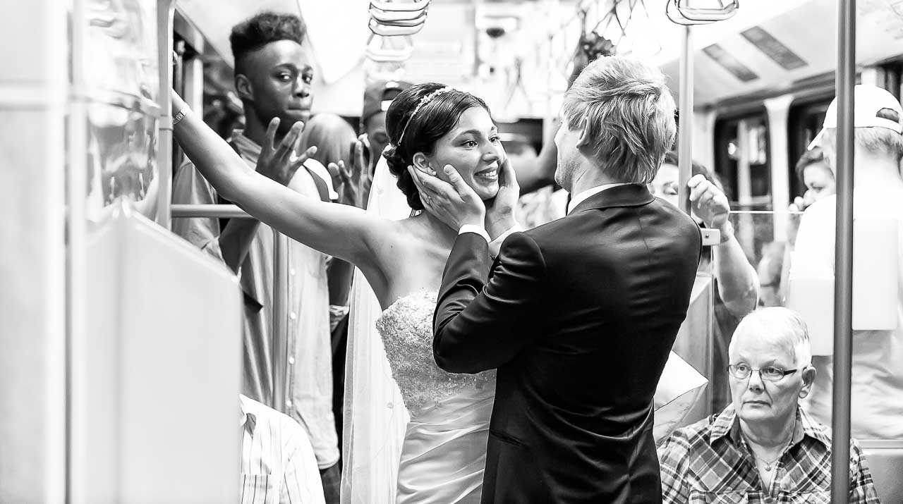 Schwarzweißfoto: Brautpaar in der U-Bahn, umgeben von Fahrgästen. Die Braut strahlt, der Bräutigam hält ihr Gesicht zärtlich. Hochzeitsfotos Hannover Hildesheim