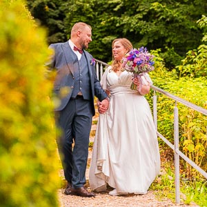 Brautpaar im Park: Braeutigam im blauen Anzug und Braut im weissen Kleid mit Blumenstrauss.