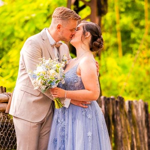 Brautpaar kuesst sich im gruenen Park, Braeutigam im hellen Anzug, Braut im blauen Kleid mit Blumenstrauss.