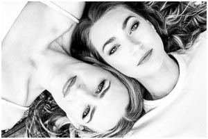 Schwarz-Weiß-Foto Pärchenfotos zweier schoener Frauen, die nah beieinander liegen. Ihre Blicke sind direkt und ausdrucksstark, ein intimer Moment der Verbundenheit.