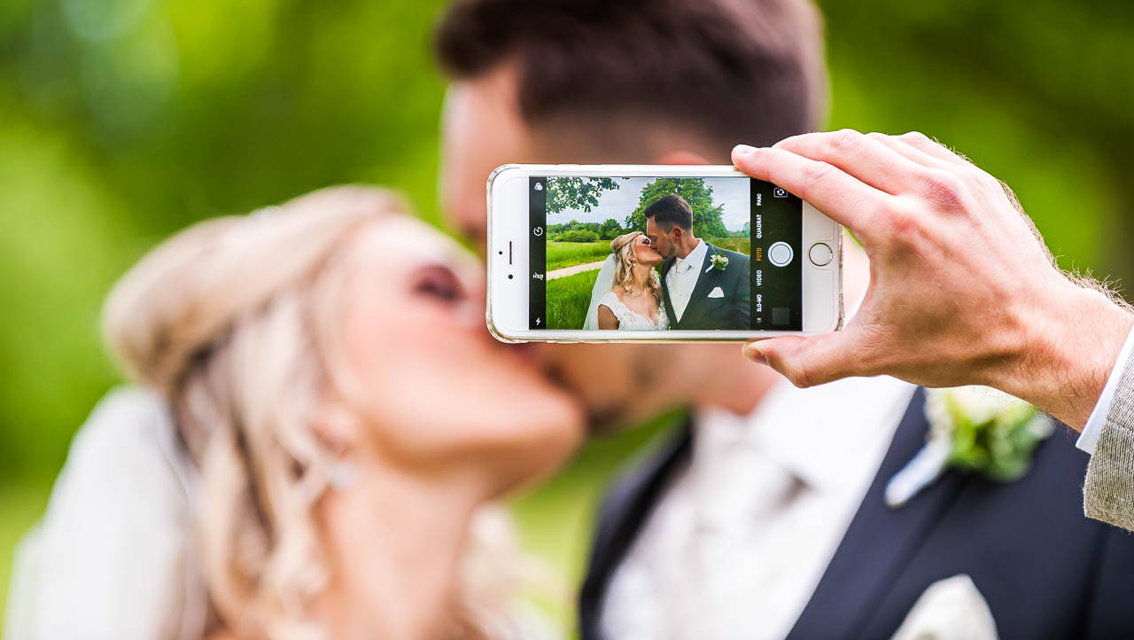 Romantisches Hochzeitsfoto: Ein Paar im innigen Kuss, festgehalten mit einem Smartphone. Schoen, wie die Liebe durch jede Aufnahme lebendig wird!