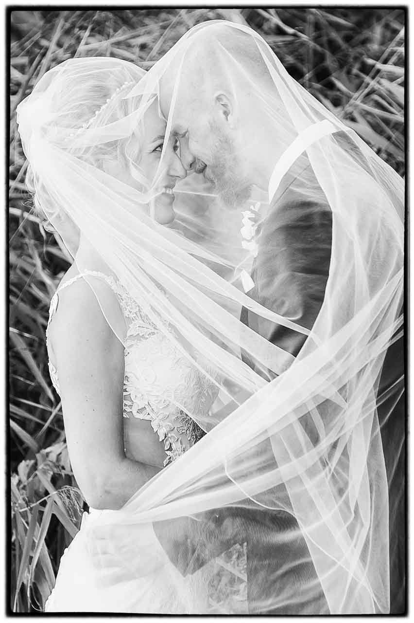 Vertraeumtes Brautpaar unter einem Schleier in Isernhagen. Sanfte Schwarz-Weiss-Aufnahme, die die Zuneigung und Intimitaet des Paares einfuehlsam zeigt.