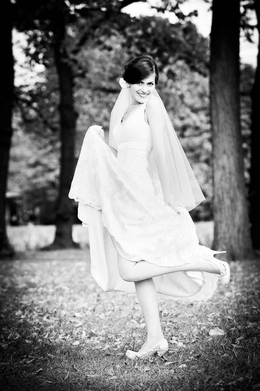 Strahlende Braut im Park von Langenhagen: Ein zauberhaftes Schwarz-Weiss-Foto zeigt eine elegante Braut mit Schleier, die ihr Kleid kokett hochhebt.